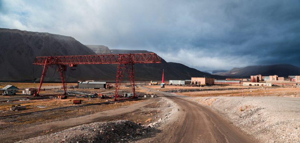 The remnants of an old mining settlement in Pyramiden, Svalbard, reveal a desolate industrial atmosphere. Rusting cranes and buildings stand against a backdrop of harsh, sweeping clouds.
