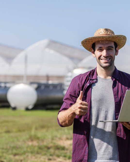 A cheerful farmer wearing a straw hat uses a laptop in front of greenhouses and agricultural equipment, symbolizing modern farming and smart agriculture technology integration.