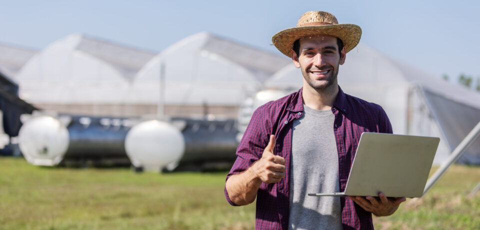A cheerful farmer wearing a straw hat uses a laptop in front of greenhouses and agricultural equipment, symbolizing modern farming and smart agriculture technology integration.