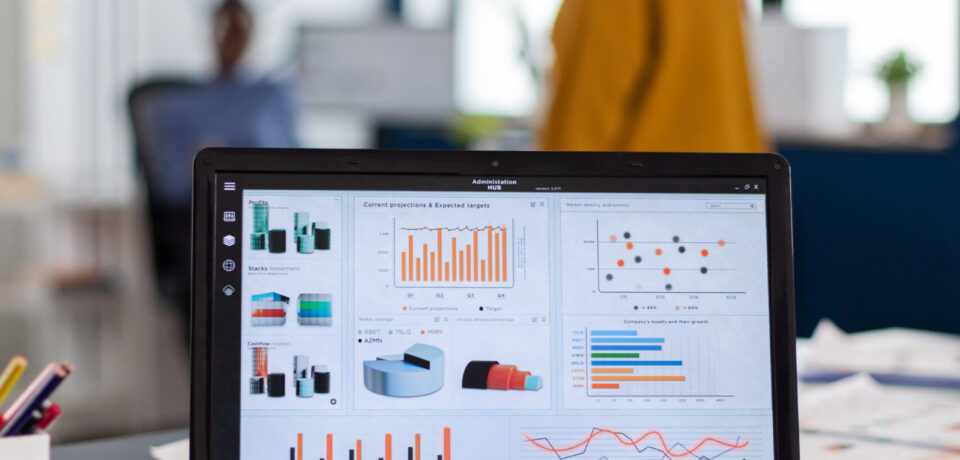 Close up of laptop with graphs, charts of marketing agency. Workplace area in business center with multi ethnic employees in it, shot of room with modern furniture and blue wall.